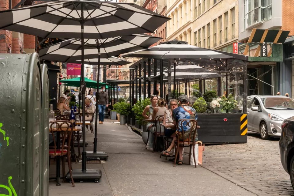 New York City sidewalk with outdoor dining patio with black and white striped umbrellas and black barriers protecting diners from traffic